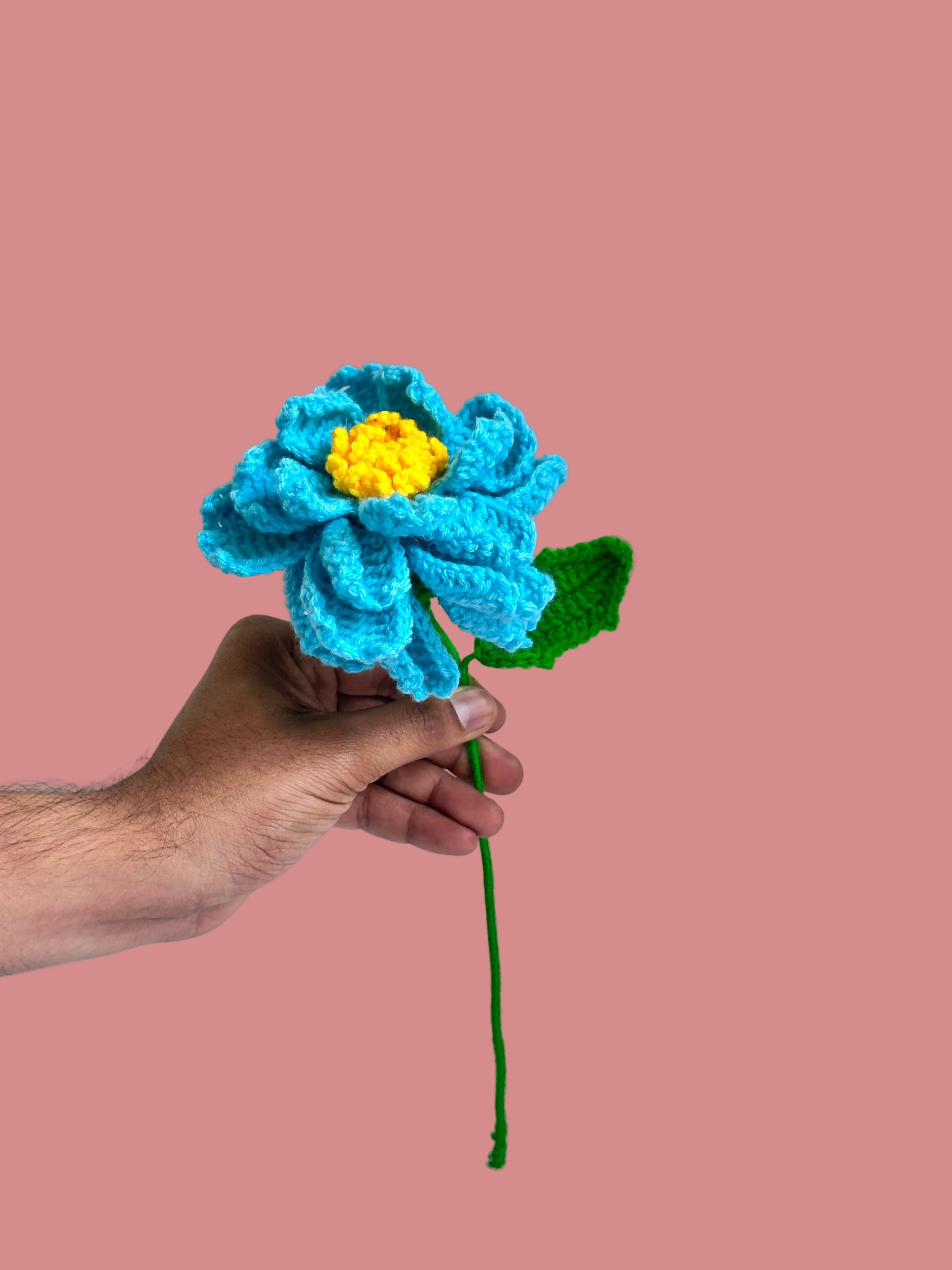 Hand holding a blue crocheted flower with a yellow center against a plain background